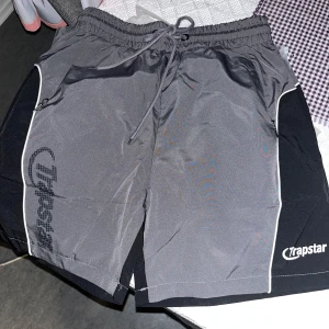 Trapstar grå och svarta shorts - Snygga grå och svarta shorts från Trapstar med vit piping längs sidorna och logga på benet. Shortsen har elastisk midja med snörning och dragkedjefickor. Perfekta för sommaren och streetwear-stil.