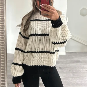 Randig stickad tröja i vitt och svart - Säljer en chunky stickad tröja i vitt med svarta ränder. Tröjan har en loose passform, breda ärmar och ribbade muddar. Perfekt för dig som gillar en avslappnad och trendig stil.