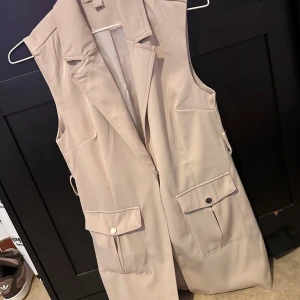 Beige lång väst från Amisu - Snygg beige väst från Amisu med klassisk krage och två stora fickor framtill med knappdetaljer. Västen är lång och har en stilren, rak passform som ger en clean look. Perfekt att slänga över en hoodie eller t-shirt för att höja outfiten.