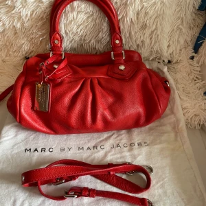 Röd skinnväska Marc by Marc Jacobs - Säljer en snygg röd handväska från Marc by Marc Jacobs i äkta skinn. Väskan har silverfärgade metalldetaljer, dubbla handtag och avtagbar axelrem. Insidan är fodrad med svartvitt mönster och har dragkedja. Perfekt för dig som vill sticka ut med en färgstark accessoar.