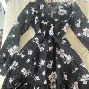 Svart blommig klänning XOXO Dreams - Supersöt svart kortklänning från XOXO Dreams med lila och vita blommor.  storlek 134/140 men KAN kanske passa en xxs eller eventuellt xs