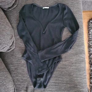 Svart v-ringad body från Zara - Svart långärmad body från Zara med djup v-ringning och ribbad struktur. Sitter tight och har knäppning i grenen. Perfekt att styla med jeans eller kjol för en clean och snygg look.