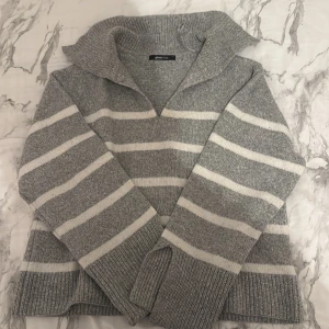 Gråvit randig stickad tröja Gina Tricot - Mysig stickad tröja från Gina Tricot i grått med vita ränder. Tröjan har en bred krage, v-ringning och långa ärmar med ribbade muddar. Perfekt för kyliga dagar och enkel att styla med jeans eller kjol.