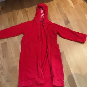 Röd badrock med huva och knytband - Mysig röd badrock med huva och knytband i midjan. Insidan av huvan har rödvita ränder. Badrocken har två stora fickor framtill och är långärmad, perfekt att slänga på sig efter duschen eller badet.