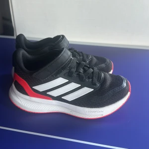 Svarta Adidas sneakers med röd detalj - Snygga svarta Adidas sneakers med vita ränder och röda detaljer på hälen. Skorna har både kardborreband och snörning för enkel påtagning. Ovandelen är i mesh och sulan är vit med Cloudfoam-dämpning för extra komfort.