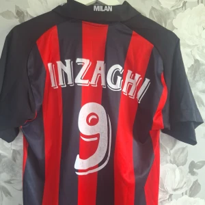 AC Milan Inzaghi #9 fotbollströja - Säljer en AC Milan fotbollströja från säsongen 1999-2000 talet med Inzaghi och nummer 9 på ryggen. Tröjan är AC Milans förstaställ för den säsongen. Tröjan har röd-svarta vertikala ränder, broderat klubbmärke och Opel-logga på bröstet. Kortärmad med krage och tillverkad i lätt polyester, perfekt för dig som älskar italiensk fotboll.