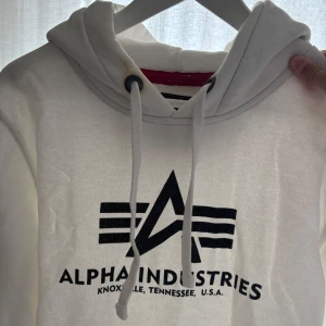 Vit hoodie Alpha Industries - Vit hoodie från Alpha Industries med svart logga framtill och klassisk känguruficka. Tröjan har huva med snörning, ribbade muddar och röd detalj vid nederkanten. Mjuk insida i fleece och långärmad modell. Perfekt för en chill och stilren look.