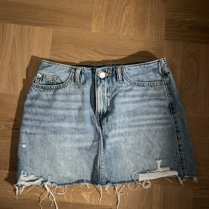 Ljusblå jeanskjol med slitna detaljer - Säljer en kort ljusblå jeanskjol med råa, fransiga kanter och slitna detaljer både fram och bak. Kjolen har klassiska fickor och bälteshällor, perfekt för en avslappnad och trendig look. Storleken är 158 men sitter exakt som en helt vanlig XS! 