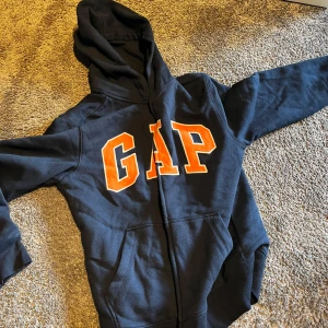 Svart GAP hoodie med orange tryck - Svart hoodie från GAP med stor orange logga framtill. Klassisk modell med huva, dragkedja och kängurufickor. Perfekt för chill dagar eller när du vill ha en avslappnad streetstyle-look.