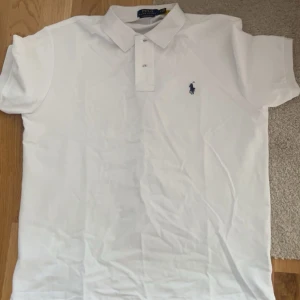 Vit Piké Polo Ralph Lauren - Klassisk vit pikétröja från Polo Ralph Lauren med korta ärmar och blå broderad logga på bröstet. Tröjan har krage och knäppning framtill, perfekt för en clean och stilren look. Tillverkad i mjuk bomull som känns skön mot huden.