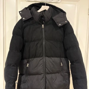 Svart pufferjacka från Brave Soul - Svart pufferjacka från Brave Soul med huva och dragkedja framtill. Jackan har quiltad design, två fickor med dragkedja och blandning av material – både matt och glansigt tyg. Perfekt för kalla dagar och riktigt snygg till streetwear.