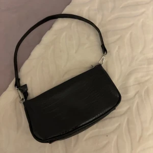 Svart axelväska  - Snygg svart axelväska med croco-mönstrad yta och silvriga metalldetaljer. Väskan har dragkedja upptill. Perfekt storlek för det viktigaste.