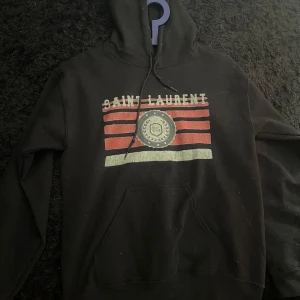 saint laurent hoodie - Svart hoodie från Saint Laurent med huva och snörning. Framsidan har ett stort tryck med röda och vita ränder samt ett emblem med SL-logga. Klassisk känguruficka och långärmad modell, perfekt för en chill och snygg streetstil.