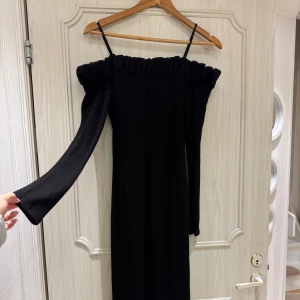 Svart offshoulder långklänning H&M - Säljer en svart långklänning från H&M med offshoulder-design och smala axelband. Klänningen har långa ärmar och är tillverkad i ribbad polyester som ger en snygg struktur. Perfekt för dig som gillar stilrena och trendiga plagg. Passar från S till M