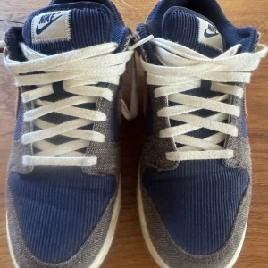 Nike Dunk Low Corduroy Blå/Brun 40 - Unika Nike Dunk Low sneakers i blå och brun manchester med vita snören och gummisula. Snygg mix av corduroy och rutigt tyg, med klassisk Nike-logga bak och på plösen. Perfekta för dig som vill sticka ut med en retrovibe och coola materialval.