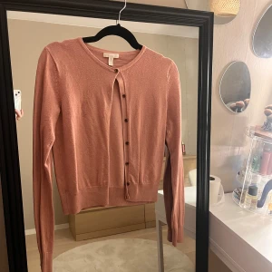 Rosa kofta med knappar från H&M - Söt ljusrosa kofta från H&M med rund halsringning och knappar framtill. Långärmad och i mjukt, tunt stickat material som passar perfekt till lager-på-lager. Enkel och stilren design som är lätt att matcha.