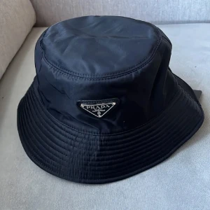 Svart bucket hat från Prada - Stilren svart bucket hat från Prada med klassisk triangulär logga i silver framtill. Hatten har rund form och bred, nedåtgående brätte. Materialet är glansig syntet som ger en lyxig vibe och passar perfekt till streetwear eller trendiga outfits.