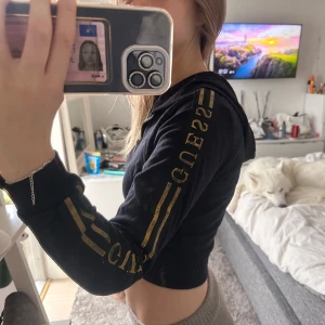 Svart croppad hoodie från Guess - Svart croppad hoodie från Guess med guldfärgade detaljer och logga längs ärmarna. Tröjan har huva, dragkedja framtill och långa ärmar. Snyggt sportig vibe och passar perfekt till streetwear. Materialet känns mjukt