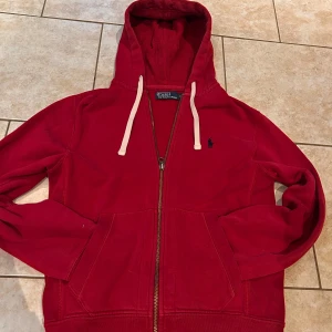 Röd hoodie från Polo Ralph Lauren - Snygg röd hoodie från Polo Ralph Lauren med klassisk blå logga på bröstet. Tröjan har huva med vita snören, dragkedja framtill och två stora fickor. Tillverkad i mjuk bomull, perfekt för chill dagar. Långärmad och med ribbade muddar för extra komfort. (Små defekter som inte märks i vardagligt bruk) perfekt för alla stilar och alla tillfällen 