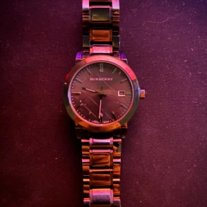 Svart Burberry klocka med länkarmband - Snygg svart Burberry klocka med rund urtavla och datumvisning. Länkarmband i metall ger en stilren och modern vibe. Swiss made med safirglas för extra hållbarhet. Perfekt accessoar för dig som gillar klassisk design med en lyxig touch.