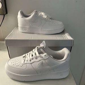 Nike Air Force 1 helvita sneakers - Klassiska Nike Air Force 1 sneakers i helvitt skinn med perforerad tå, vit sula och snörning. Snygg och tidlös modell med låg profil och diskret Nike-logga på sidan. Perfekta för dig som gillar cleana och fräscha sneakers.