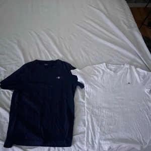 Två GANT t-shirts marinblå & vit - Säljer två stilrena GANT t-shirts i storlek 146/152. Ena är marinblå och den andra vit, båda med klassisk GANT-logga broderad på bröstet. T-shirtsen har rund hals och korta ärmar, tillverkade i mjuk bomull. Perfekt för dig som gillar enkel och snygg stil. Nypris 400 för båda. Hör av er vid fungeringar!