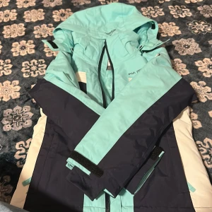 Turkos McKinley skidjacka strl 120 - Säljer en fräsch turkos och mörkblå skidjacka från McKinley, modell Brenda JR. Jackan har huva, dragkedja och praktiska fickor. Materialet är slitstark polyester och insidan är glansig. Perfekt för vinter och skidåkning. Snygg färgkombination med vita detaljer. Helt oanvänd köpt för 799kr.