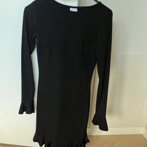 Svart volangklänning från Gina Tricot - Supersnygg svart klänning från Gina Tricot i storlek XS. Modellen är figurnära med långa ärmar och volangdetaljer vid ärmslut och nederkant. Klänningen har en enkel, stilren design och är perfekt för dig som gillar en chic look med lite edge.