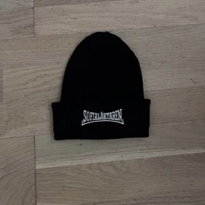 Svart ribbad mössa Sofialäktaren - Svart ribbstickad mössa med uppvikt kant och vit broderad text 'Sofialäktaren' framtill. Perfekt accessoar för att visa support och hålla värmen. Enkel och stilren design som passar till det mesta.