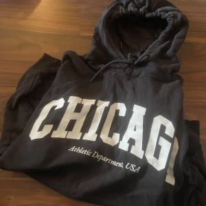 Svart hoodie med CHICAGO-tryck - Svart hoodie med stor vit CHICAGO-text på bröstet och snörning i huvan. Mjuk bomullskänsla och klassisk streetwear-look, perfekt för dig som gillar sportiga vibes och college-stil. Snygg att matcha med jeans eller joggers.