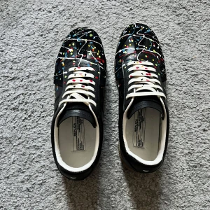 Maison Margiela sneakers med färgstänk - Unika svarta sneakers från Maison Margiela med coola färgstänk i rött, blått, gult och vitt. Skorna har vita snören, beige gummisula och är tillverkade i skinn. Perfekt för dig som vill sticka ut med en konstnärlig vibe.