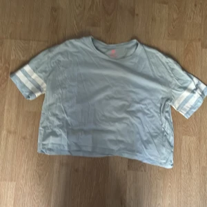 Ljusblå t-shirt med ränder från H&M - Ljusblå t-shirt från H&M i ekologisk bomull. Modellen är loose fit med korta ärmar och två vita ränder på varje ärm för en sportig vibe. Perfekt för dig som gillar enkel och chill stil.