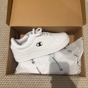Vita sneakers från Champion - Fräscha vita sneakers från Champion med klassisk design och svart logga på sidan. Skorna har snörning och är tillverkade i syntetmaterial med en platt sula och rund tå. Perfekta för en clean och sportig look.