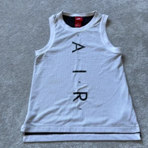 Vit linnetopp från Nike AIR - Säljer en sportig vit linnetopp från Nike med svart text 'AIR' vertikalt framtill. Toppen är i meshmaterial som andas och har ärmlös design, perfekt för träning eller chill dagar. Klassisk Nike-stil med rund hals och snygga detaljer.
