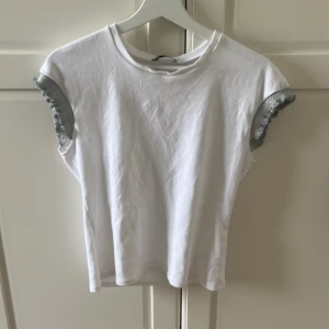 Vit topp med glittriga volangärmar - Snygg vit topp med korta ärmar och glittriga silvervolanger vid axlarna. Enkel rund halsringning och rak passform. Perfekt för dig som vill ha något extra till outfiten. Gjord i mjuk bomull med detaljer i glittrigt material. Storlek L men passar mig bra som har M.