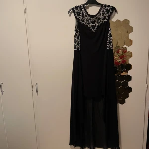 Svart långklänning med pärldetaljer - Säljer en svart långklänning med ärmlös design och transparent nederdel. Överdelen är dekorerad med glittrande pärlor i ett snyggt mönster som ger en lyxig känsla. Perfekt för bal eller fest. Klänningen har rund halsringning och faller snyggt längs kroppen.