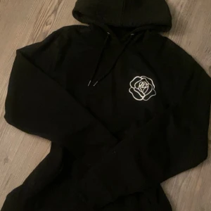 Svart hoodie med ros och text - Svart hoodie med huva och snörning från Trycklagret. Helt oanvänd och jättebra skick. Mjuk och gosig.