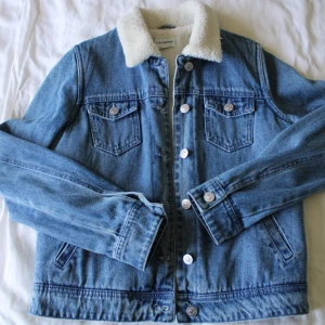 Blå jeansjacka med teddykrage - Snygg blå jeansjacka från Denim Prime med vit teddykrage. Klassisk modell med två bröstfickor, silverfärgade knappar och långa ärmar. Perfekt för dig som vill ha en cool och tidlös look med extra mysig känsla tack vare den mjuka kragen. Tagg är borttagen men den är aldrig buren, enbart prövad. Blev tyvärr mer populärt med hoodie och vanlig jeansjacka för mig så denna kom aldrig till användning. Ingen fordring i ärmar! 