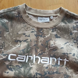 Carhartt camouflage sweatshirt - Säljer en beige och grå camouflage-mönstrad sweatshirt från Carhartt med stor vit broderad logga på bröstet. Tröjan har rund hals, ribbade muddar och är i mjuk bomull. Perfekt för dig som gillar streetwear och vill sticka ut. Sitter perfekt, materialet är mycket behaglig vid beröring. Mer information, bilder och prisdiskutering i dm. Äkta