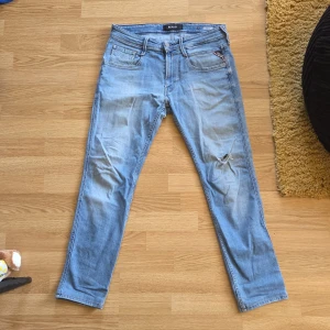 Replay Anbass ljusblå jeans W31 L32 - Säljer ett par Replay Anbass jeans i ljusblå tvätt med klassisk femficksdesign och diskret Replay-logga på myntfickan. Jeansen har en slim passform och ett litet hål på höger knä för en avslappnad look. Perfekta till sneakers och hoodie.
