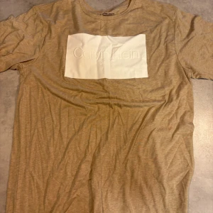 Beige t-shirt Calvin Klein Jeans XL -  Calvin Klein Jeans T-shirt beige i storlek XL. Framsidan har en vit, upphöjd logga som ger en clean och modern vibe. T-shirten är gjord i mjuk bomull och polyester, har rund hals och korta ärmar. Perfekt för dig som gillar stilrena och enkla plagg.