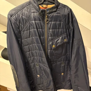 Barbour quiltad jacka Steve McQueen L - Snygg quiltad jacka från Barbour International Steve McQueen-kollektionen. Mörkblå med orange detaljer, dragkedja framtill och flera fickor med tryckknappar. Insidan har ett coolt tryck med brittiska och amerikanska flaggan. Perfekt för dig som gillar klassisk stil med edge.