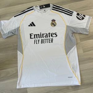 25/26 Real Madrid hemmatröja Adidas vit (finns i L och M) - Snygg Real Madrid hemmatröja från Adidas med klassisk vit bas, svarta detaljer och gula linjer. Tröjan har klubbmärke på bröstet, sponsortryck och svarta ränder på axlarna. Tillverkad i lätt och ventilerande material, perfekt för fotbollsträning eller match.