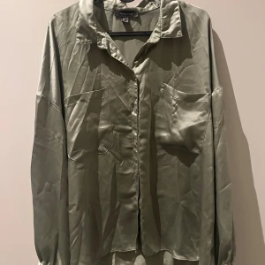 Olivgrön satinblus från Primark - Snygg olivgrön blus från Primark i satinliknande material. Modellen har långa ärmar med resår vid ärmslut, två stora bröstfickor och klassisk krage. Knappar framtill ger en clean look. Perfekt för dig som gillar stilrena och trendiga plagg.