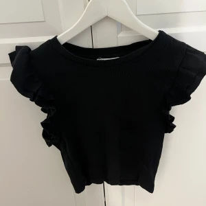 Svart ribbad topp med volangärmar - Snygg svart ribbad topp med volangdetaljer på axlarna. Modellen är ärmlös och har en rund halsringning. Perfekt att styla med jeans eller kjol för en trendig look. Säljer då den inte passat mig längre.💞