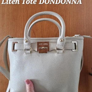 Beige handväska från Don Donna - Snygg liten beige handväska från Don Donna med två handtag och axelrem. Väskan har guldfärgade metalldetaljer, dragkedja upptill och ett extra fack med dragkedja på baksidan. Tillverkad i syntetmaterial med stilren och klassisk design.