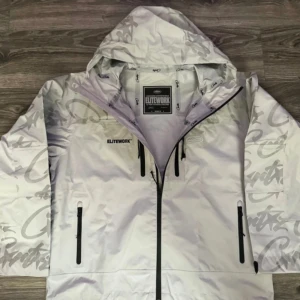 Grå cortiez elitework vindjacka - Snygg grå vindjacka från cortiez med grått graffiti-inspirerat mönster. Jackan har huva, svarta dragkedjor och flera fickor framtill. Perfekt för dig som gillar streetstyle och vill sticka ut med coola detaljer.