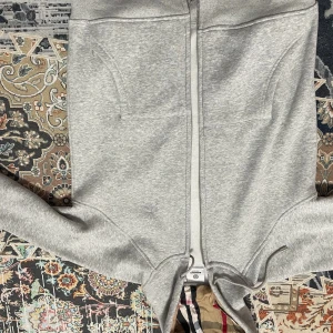 Grå hoodie från Burberry med dragkedja - Grå hoodie från Burberry med klassiskt rutigt foder i huvan, hel dragkedja och snörning. Tröjan har två stora fickor framtill och diskret broderad logga på bröstet. Tillverkad i mjuk bomullsmix, perfekt för chill dagar. 