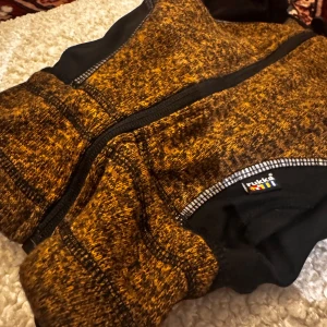 Rukka hundkläder i svart och brunt  - Säljer ett par täckbyxor från Rukka i svart och brunt melerat tyg. Byxorna har dragkedja framtill, elastiska partier och är tillverkade i polyester med lite stretch. Perfekta för kalla dagar när du vill hålla dig varm och bekväm.