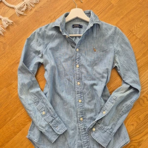 Ljusblå skjorta från Polo Ralph Lauren - Säljer en klassisk ljusblå skjorta från Polo Ralph Lauren i 100% bomull. Skjortan har lång ärm, knappar framtill och den ikoniska broderade loggan på bröstet. Perfekt för en clean och avslappnad look.